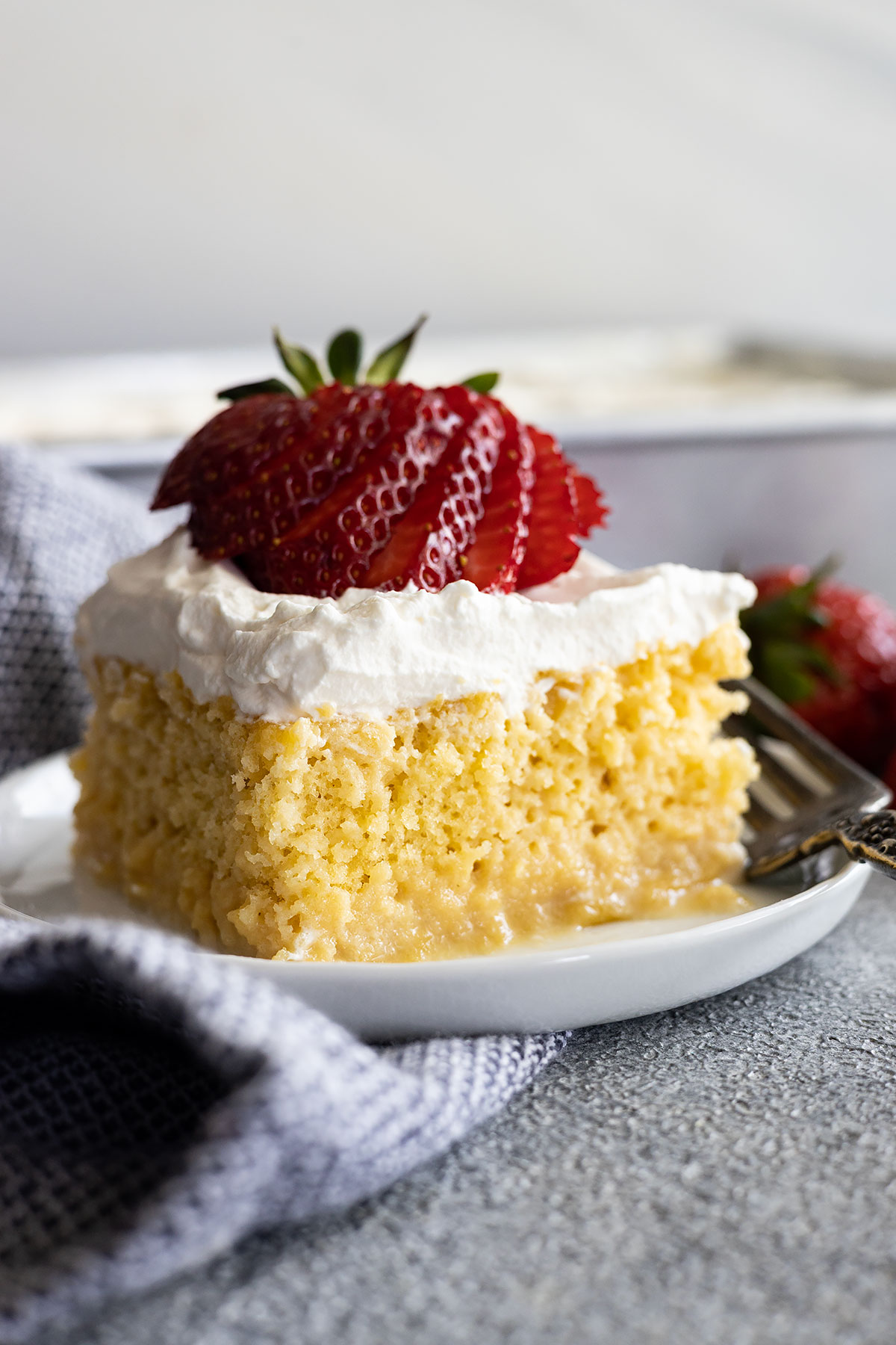 A slice of tres leches cake on a white plate topped with a sliced strawberry fanned out.
