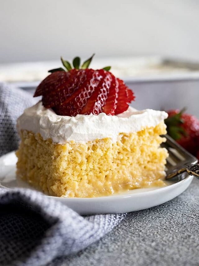 A piece of tres leches cake on a small plate topped with a fanned out strawberry.