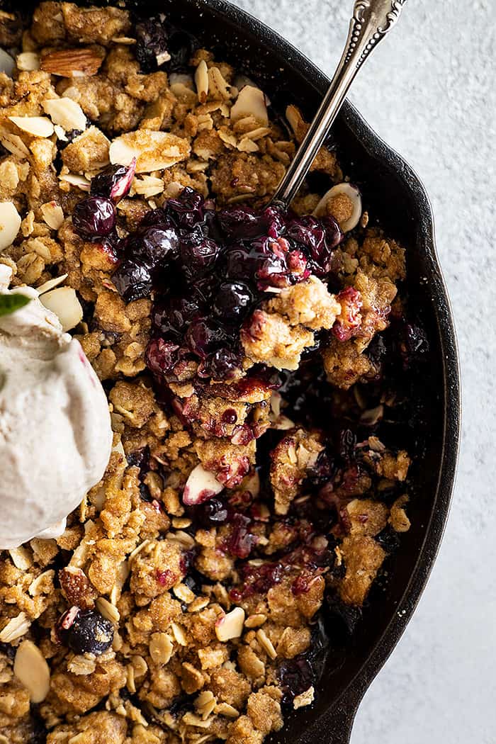 Close up of blueberry crisp with a spoon in it. Topped with a scoop of ice cream. 