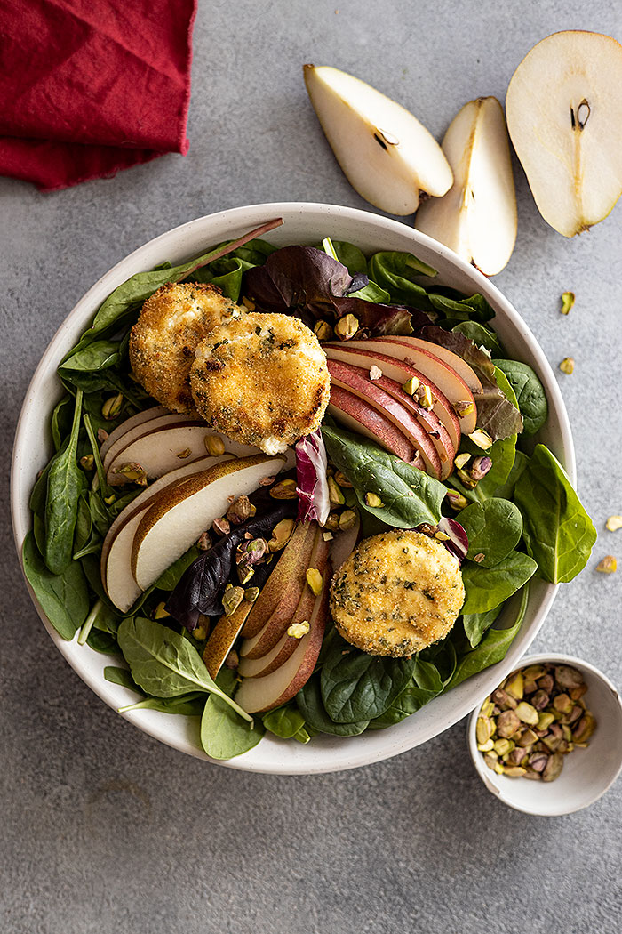 Overhead view of salad with sliced pears and goat cheese medallions and sprinkled with chopped pistachios. 