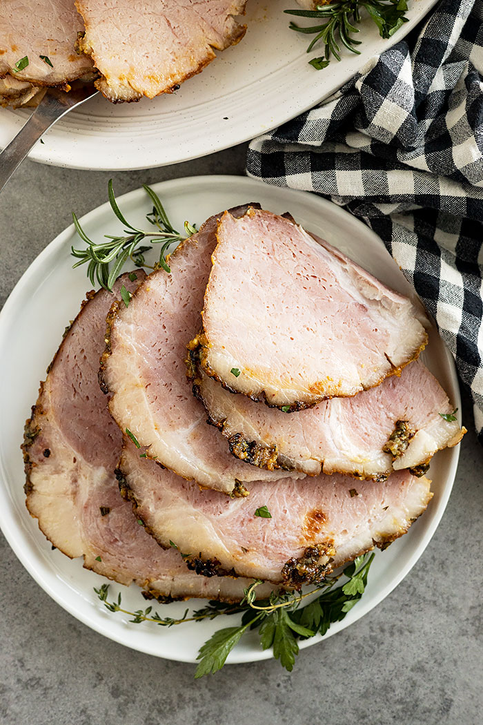 Overhead view of several slices of mustard herb ham on a plate garnished with fresh herbs.