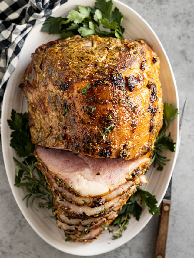 Overhead shot of ham on a white plate garnished with herbs. Part of the ham is sliced.