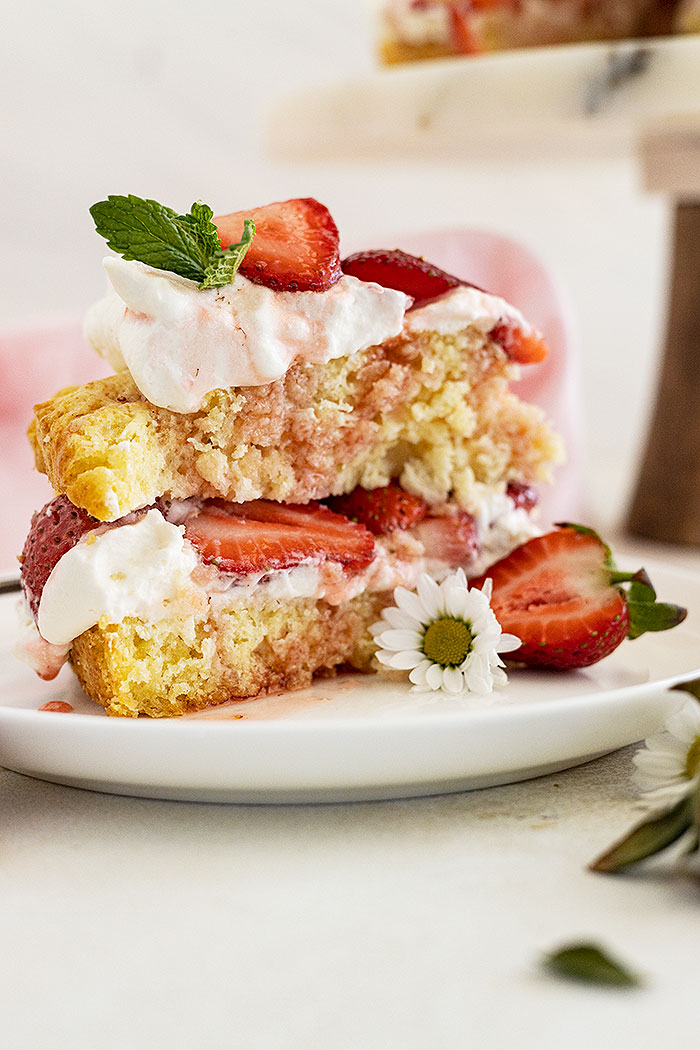 A slice of strawberry shortcake cake on a white plate and garnished with mint and little white flowers.