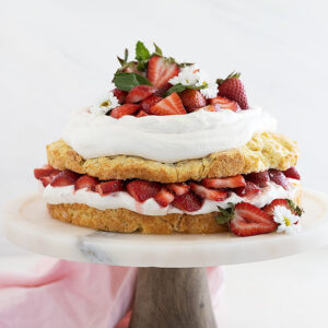 Straight on shot of strawberry shortcake cake on a cake stand and garnished with mint and strawberries cut in half.