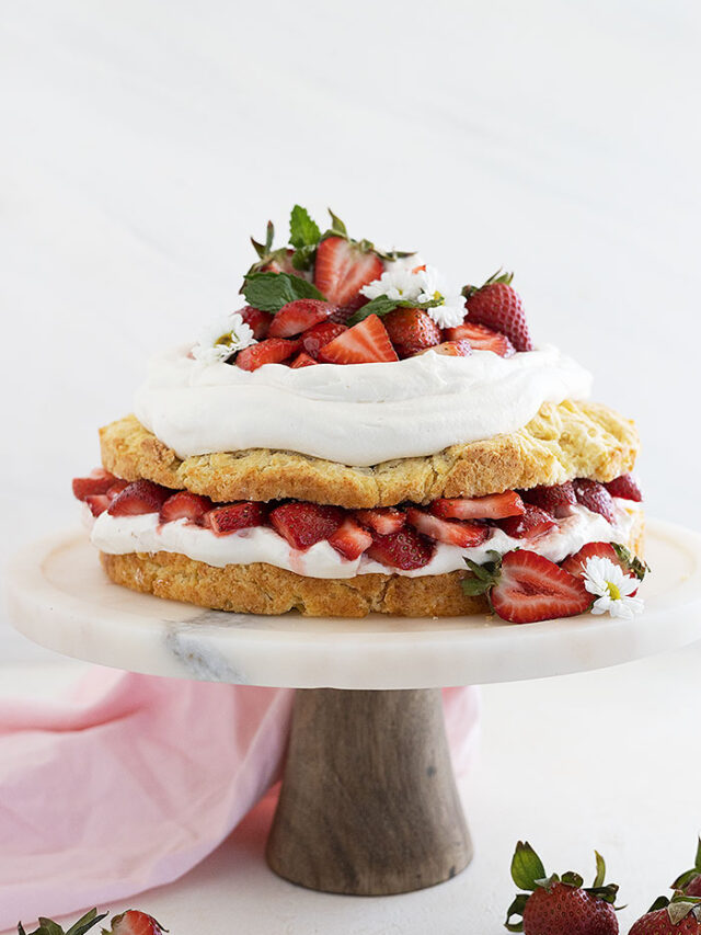 Straight on shot of strawberry shortcake cake on a cake stand and garnished with mint and strawberries cut in half.