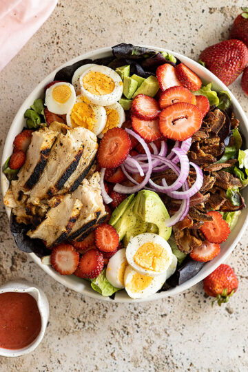 Overhead view of a large bowl of strawberry cobb salad with dressing off to the side.