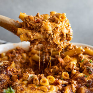 Pulling up a large spoonful of baked ziti from a white casserole dish.