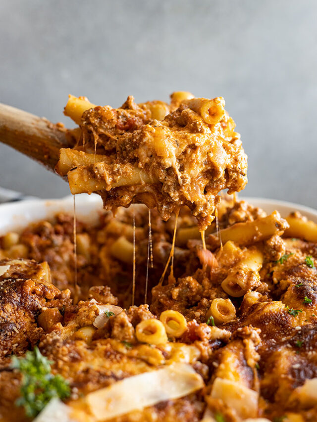 Pulling up a large spoonful of baked ziti from a white casserole dish.