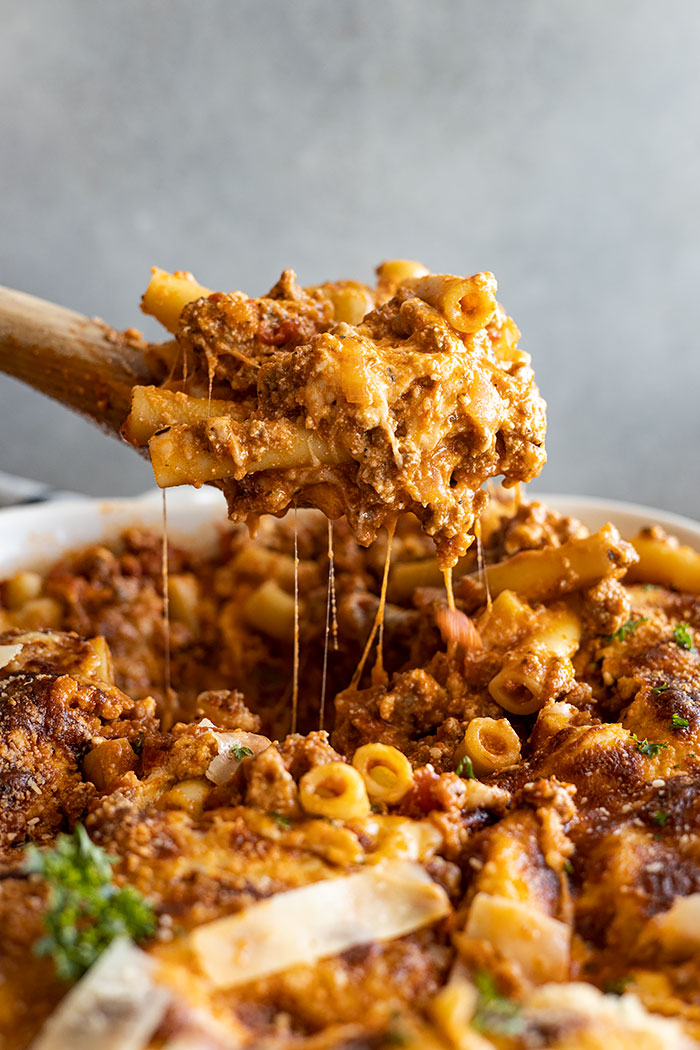 Pulling up a large spoonful of baked ziti from a white casserole dish. 