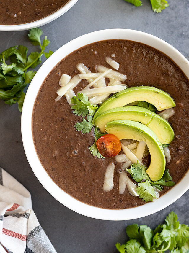 Overhead view of black bean soup in a white bowl garnished with avocado, cheese, cilantro, and tomato.