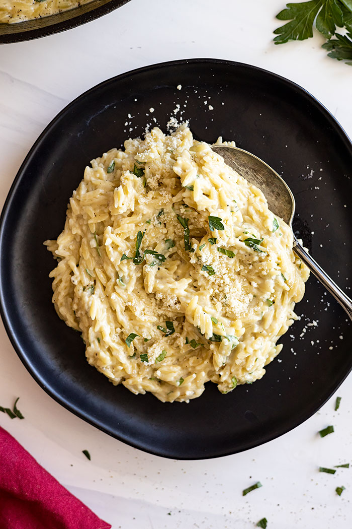 Orzo served on a black plate with a spoon ready to take a bite. Garnished with fresh parsley , parmesan cheese, and black pepper. 