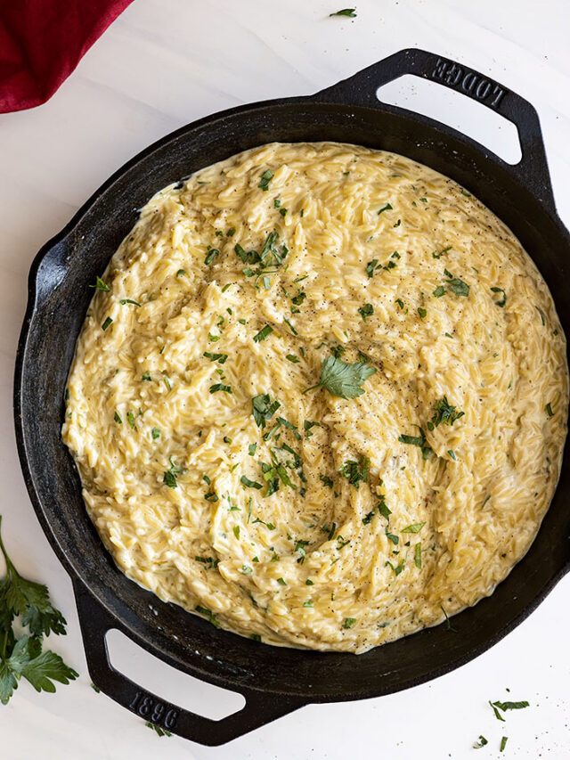 Overhead view of parmesan orzo in a large black skillet. Garnished with fresh parsley and black pepper.