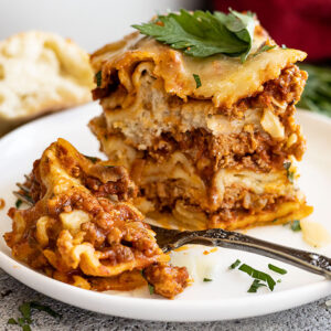 A tall slice of lasagna on a white plate with a bite taken out. Garnished with fresh parsley and parmesan cheese.