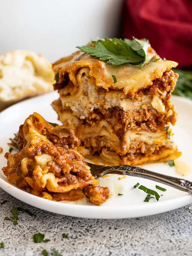 A tall slice of lasagna on a white plate with a bite taken out. Garnished with fresh parsley and parmesan cheese.
