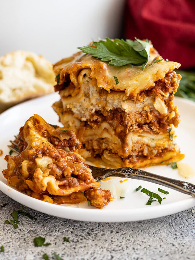 A tall slice of lasagna on a white plate with a bite taken out. Garnished with fresh parsley and parmesan cheese.