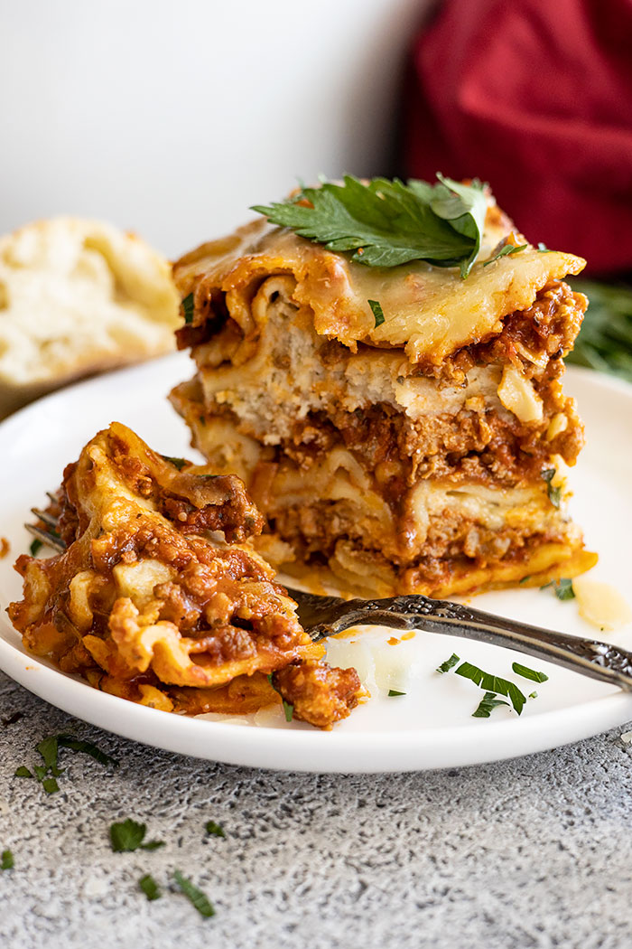 A tall slice of lasagna on a white plate with a bite taken out. Garnished with fresh parsley and parmesan cheese. 