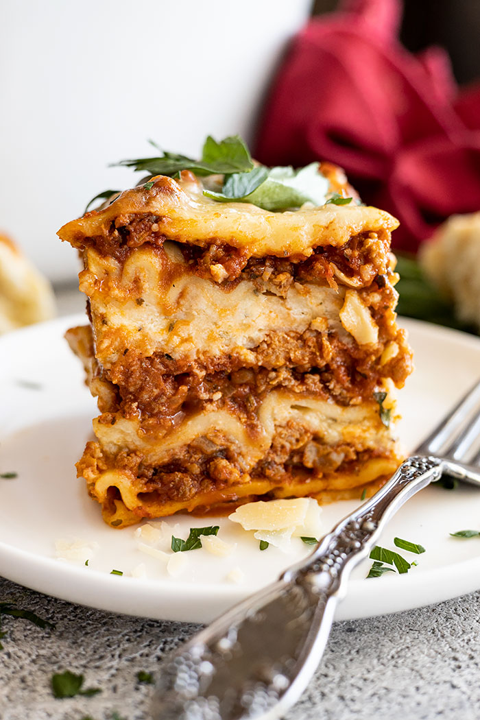 A piece of lasagna on a white plate showing all the yummy layers. Garnished with parmesan cheese and fresh parsley. 