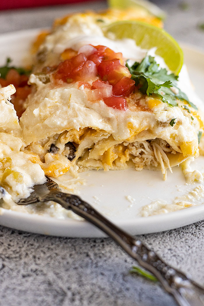 White chicken enchiladas on a white plate with a fork full of enchilada off to the side. Can see the yummy chicken interior of the enchilada.