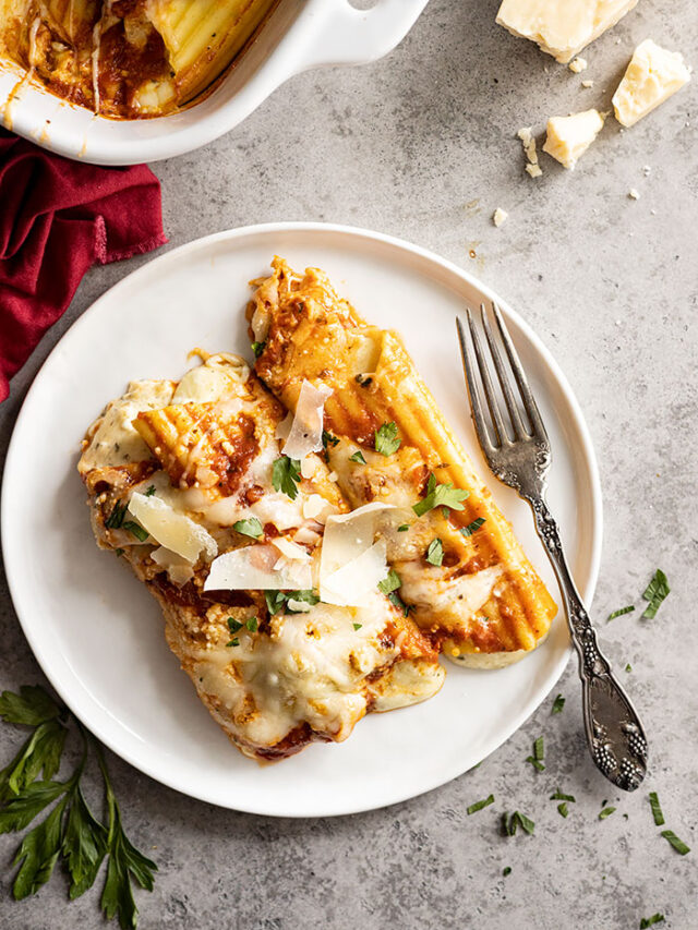 Overhead view of manicotti on a white plate. Garnished with fresh parsley and parmesan cheese. Fork and red napkin off to the side.