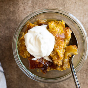 Overhead view of pumpkin bread pudding in a glass bowl drizzled with caramel sauce and a dollop of whipped cream.