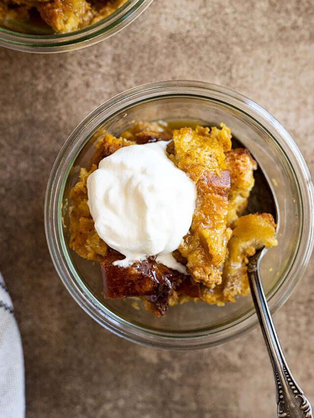 Overhead view of pumpkin bread pudding in a glass bowl drizzled with caramel sauce and a dollop of whipped cream.