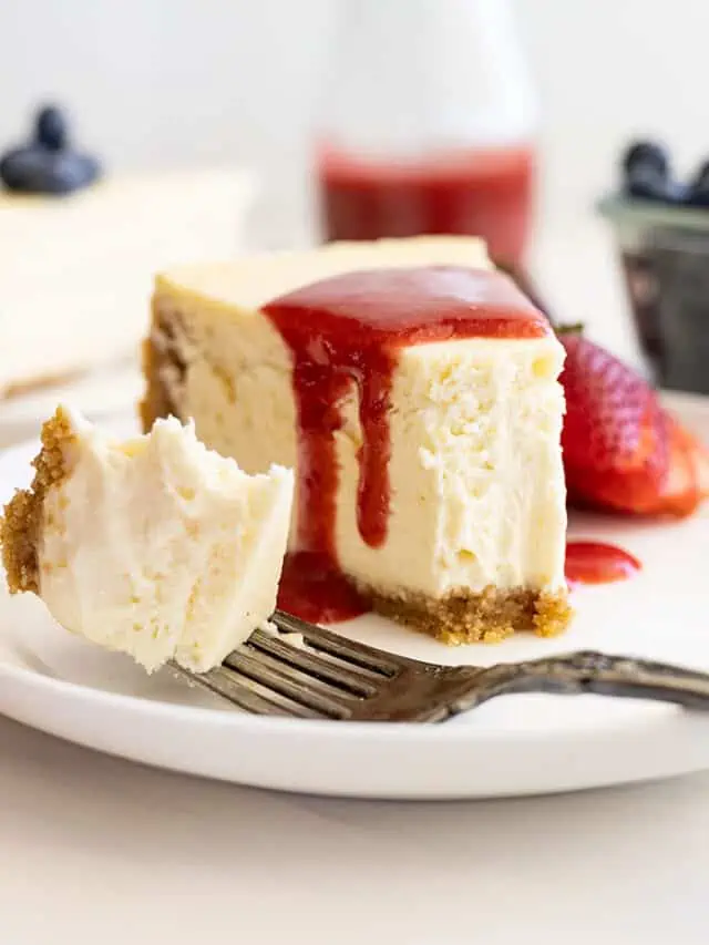 Straight on view of vanilla cheesecake on a white plate, strawberry sauce over the top, and a bite taken out showing how creamy and smooth it is.