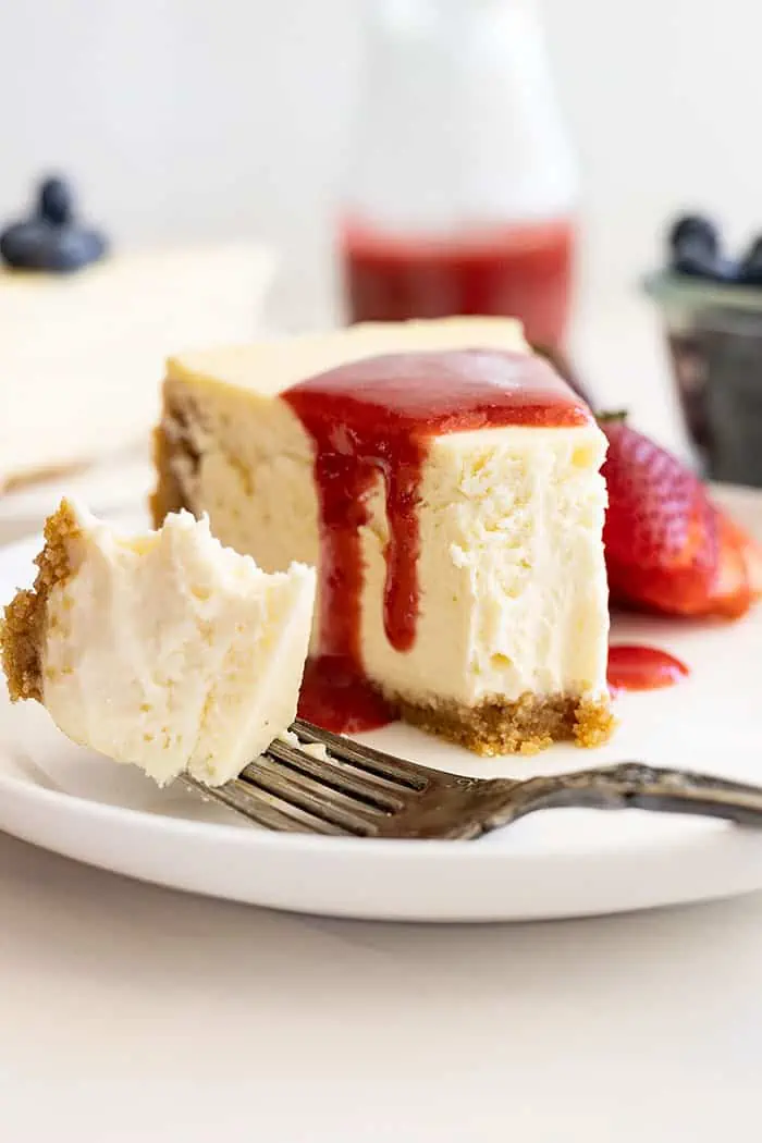 Straight on view of vanilla cheesecake on a white plate, strawberry sauce over the top, and a bite taken out showing how creamy and smooth it is. 