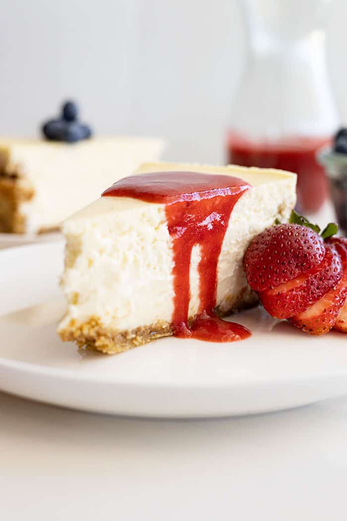 Straight on view of a slice of cheesecake with strawberry sauce spilling over the edge. 
