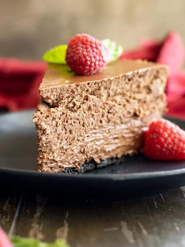 Slice of chocolate cheesecake on a black plate with a raspberry and mint for garnish.