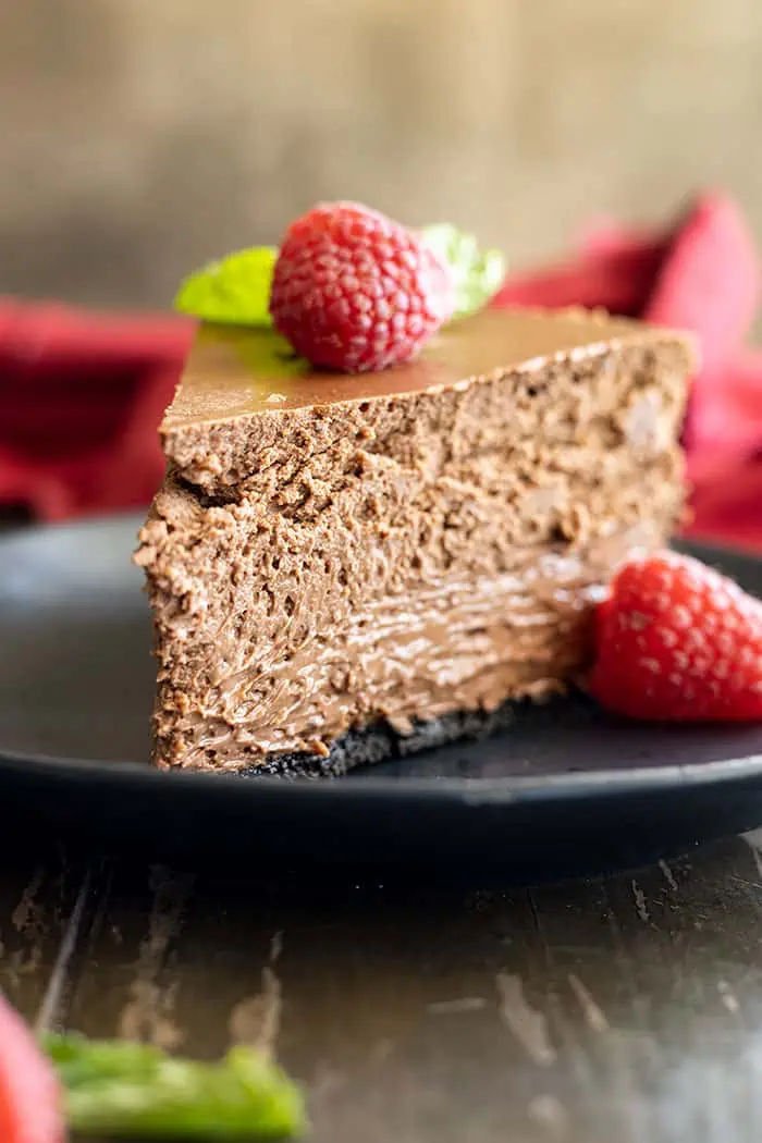 Slice of chocolate cheesecake on a black plate with a raspberry and mint for garnish. 