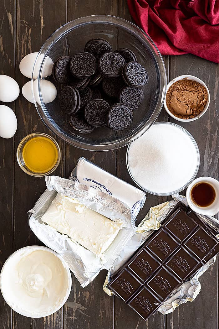Ingredients to make cheesecake: oreos, butter, sugar, cocoa powder, eggs, chocolate, cream cheese, sour cream, and vanilla.
