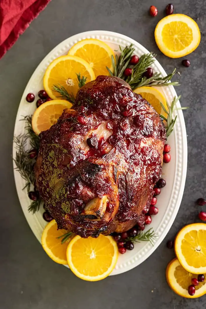 Overhead view of glazed ham on a white plate garnished with orange slices, rosemary, and fresh cranberries.