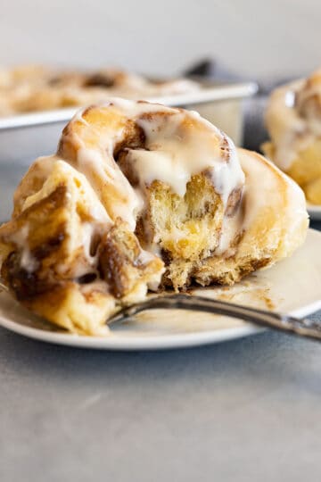 Cinnamon roll on a white plate with a bite on a fork to the side.