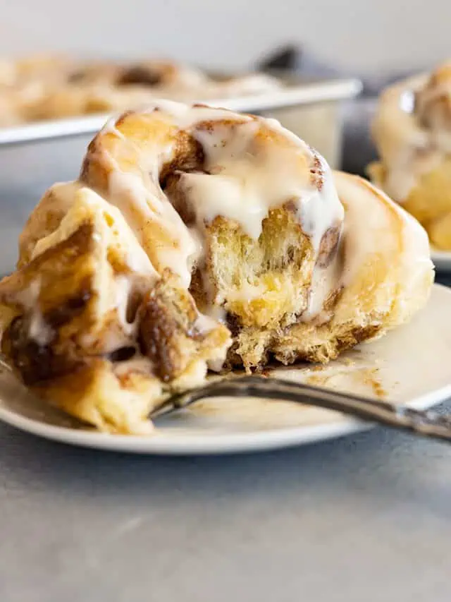 Cinnamon roll on a white plate with a bite on a fork to the side.