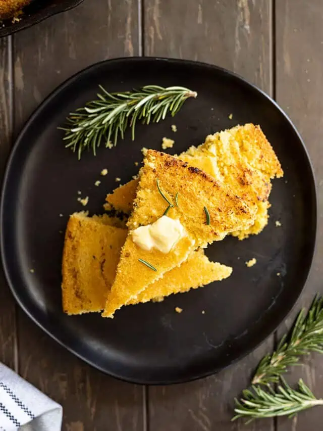 Overhead view of slices stacked on a black plate with melting butter on the top. Rosemary to the side.