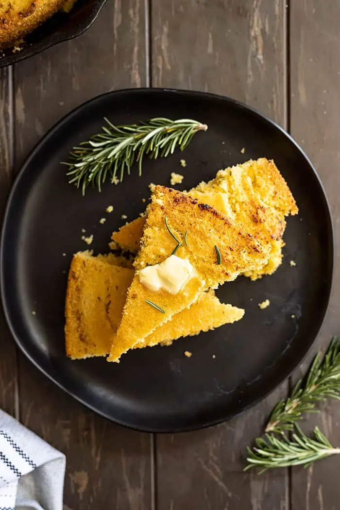 Overhead view of slices stacked on a black plate with melting butter on the top. Rosemary to the side. 