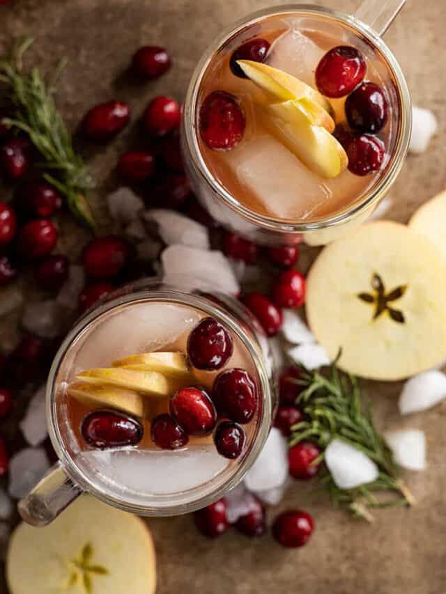 Overhead view of two mugs of apple cranberry ginger beer garnished and surrounded by apple slices, cranberries, crushed ice, and rosemary springs.