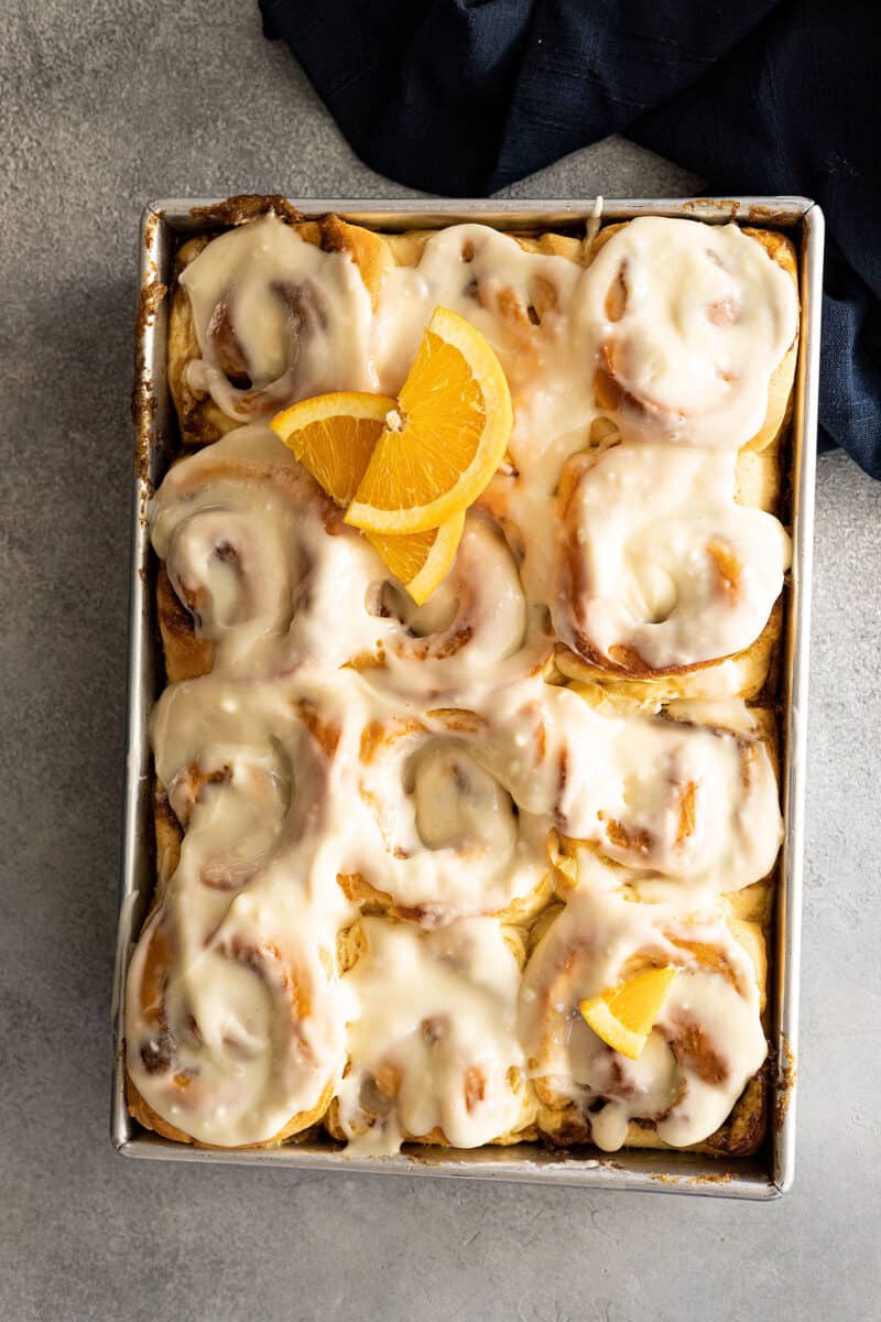 Overhead view of orange cinnamon rolls topped with cream cheese frosting and a couple orange slices for garnish. 
