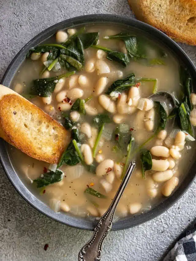 Overhead view of a bowl of tuscan bean soup in a dark bowl. Crusty bread off to the side and a spoon in the soup.