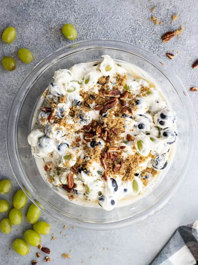 Overhead view of creamy grape salad in a bowl garnished with chopped nuts and brown sugar. Grapes and nuts sprinkled around the bowl.