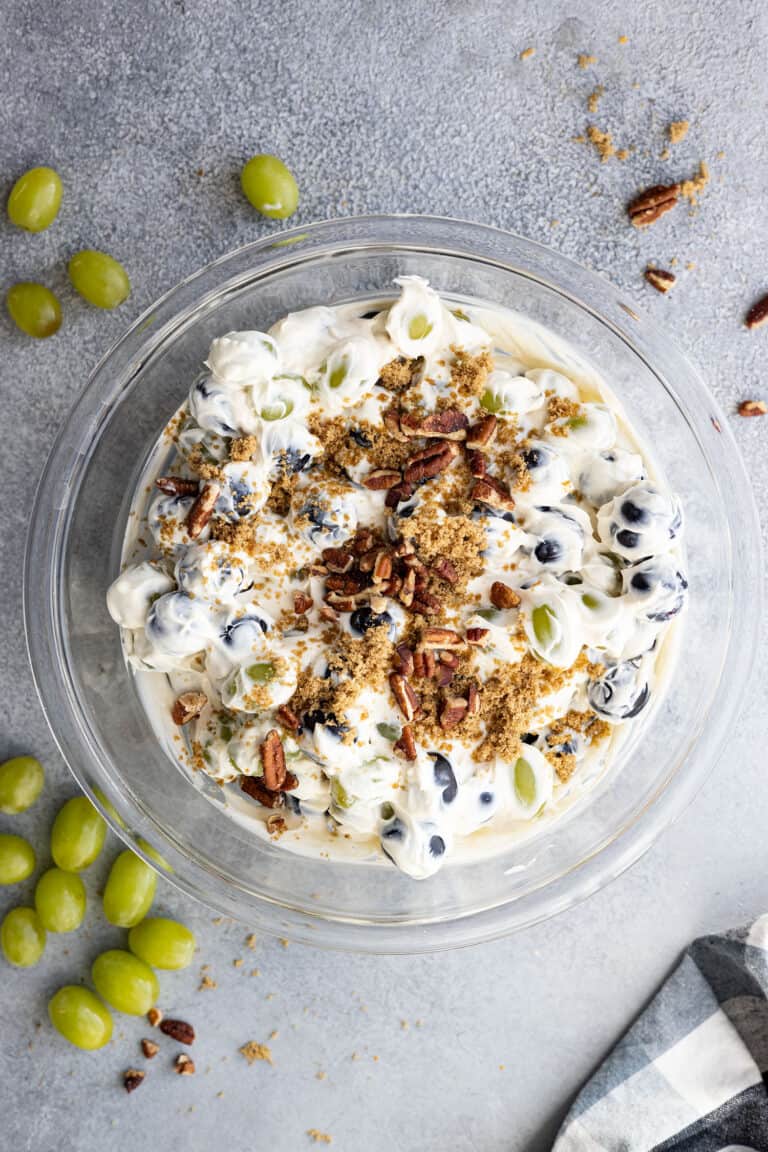 Overhead view of creamy grape salad in a bowl garnished with chopped nuts and brown sugar. Grapes and nuts sprinkled around the bowl.
