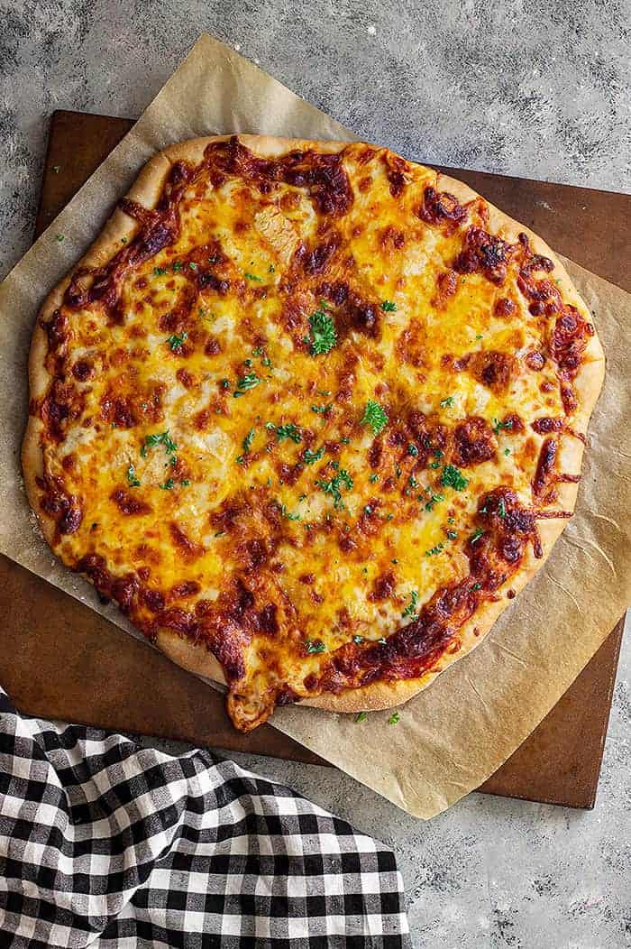 overhead: homemade cheese pizza on parchment paper on a wooden cutting board with a black and white checkered towel to the left