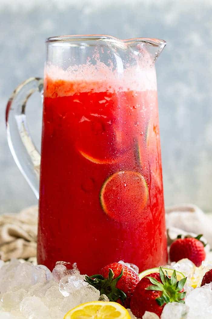 a tall pitcher of lemonade margarita on ice with strawberries and limes 