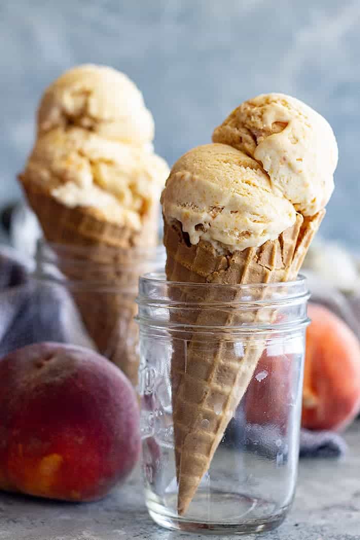 side view: ice cream cones topped with scoops of peach ice cream in ball jars. Fresh peaches are shown off to the side