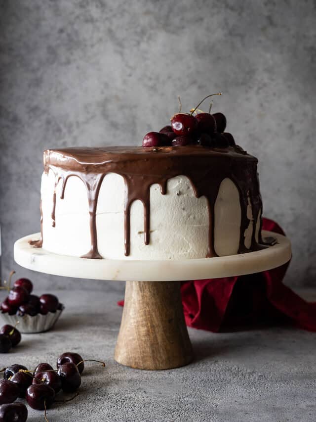 Black forest cake on a stand topped with ganache and fresh cherries.