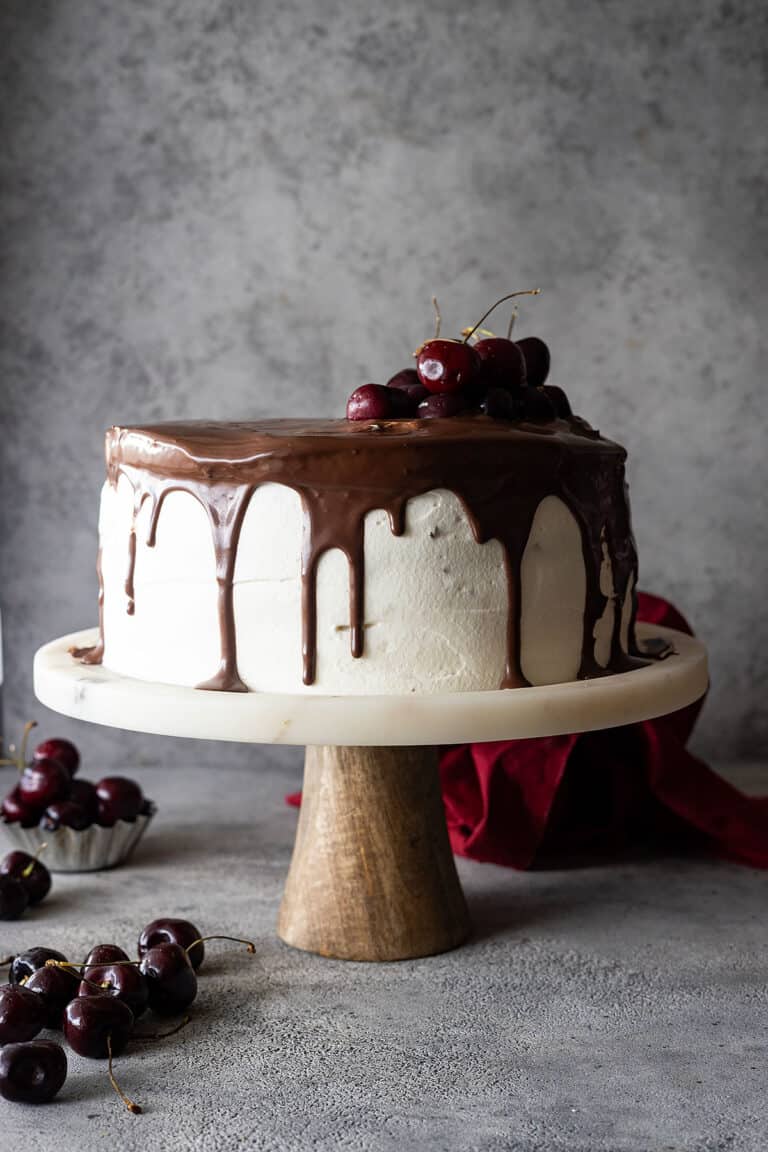 Black forest cake on a stand topped with ganache and fresh cherries.