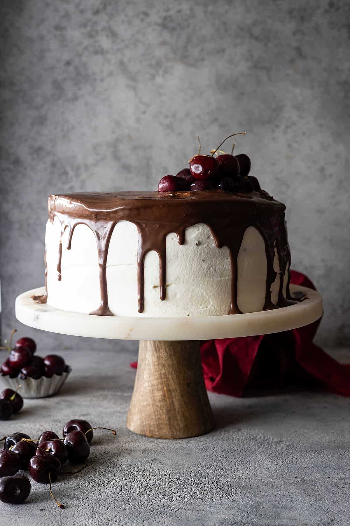 Black forest cake on a stand topped with ganache and fresh cherries.