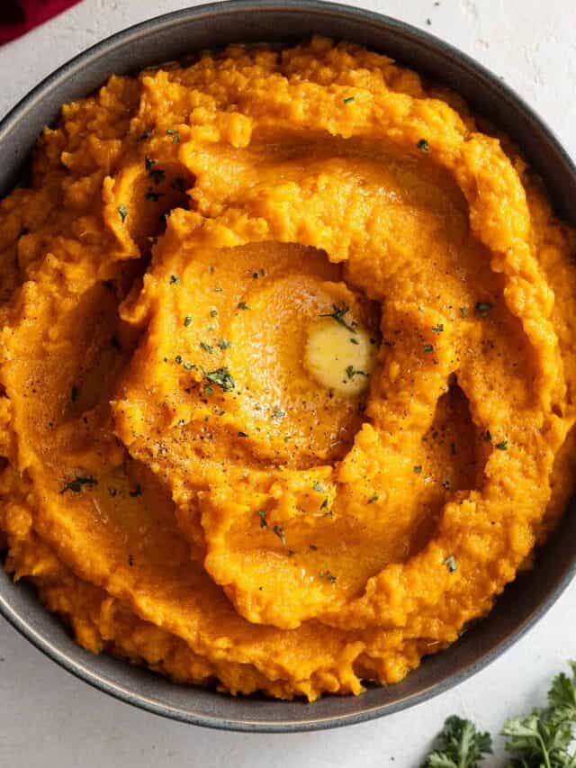 Overhead view of mashed sweet potatoes in a dark colored bowl. Garnished with black pepper, parsley, and melted butter.