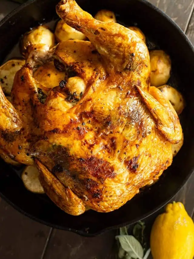 Overhead view of roasted chicken fresh from the oven. Lemon and fresh herbs off to the side.