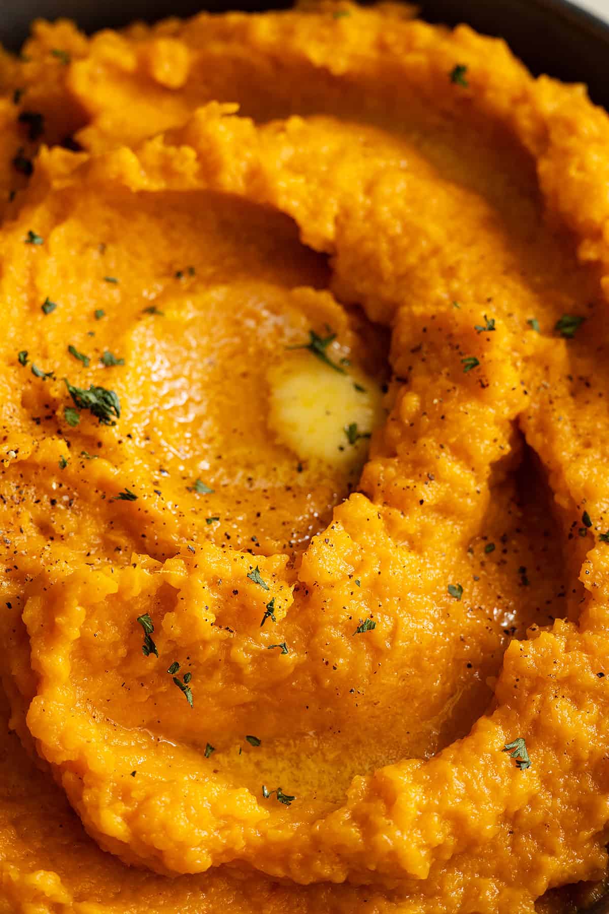Close up of mashed sweet potatoes with pools of melted butter, fresh black pepper, and parsley. 